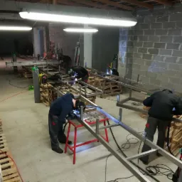 Warsztat z pracownikami spawającymi metalowe konstrukcje na paletach, widoczne butle z gazem i narzędzia spawalnicze, surowe wnętrze z betonowymi ścianami.