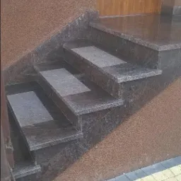 Zewnętrzne schody wejściowe z ciemnego kamienia z jasnymi wstawkami, prowadzące do drewnianych drzwi wejściowych, obok fragmentu brukowanej ścieżki.