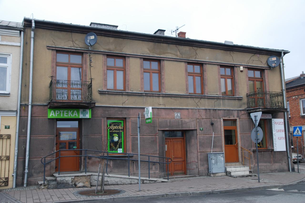 Dwukondygnacyjny budynek z brązową elewacją i drewnianą stolarką okienną, z balkonami z kutą balustradą. Widoczne pęknięcia na elewacji i szyld apteki.