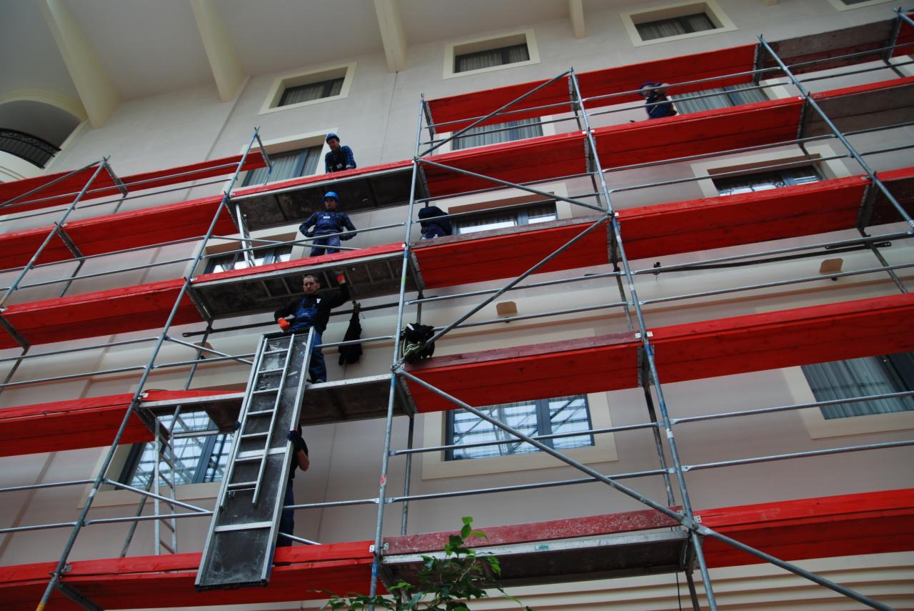 Praca na rusztowaniu: ekipa w roboczych strojach przy elewacji budynku z oknami. Czerwone deski rusztowania, widok z dołu, lekki kąt.