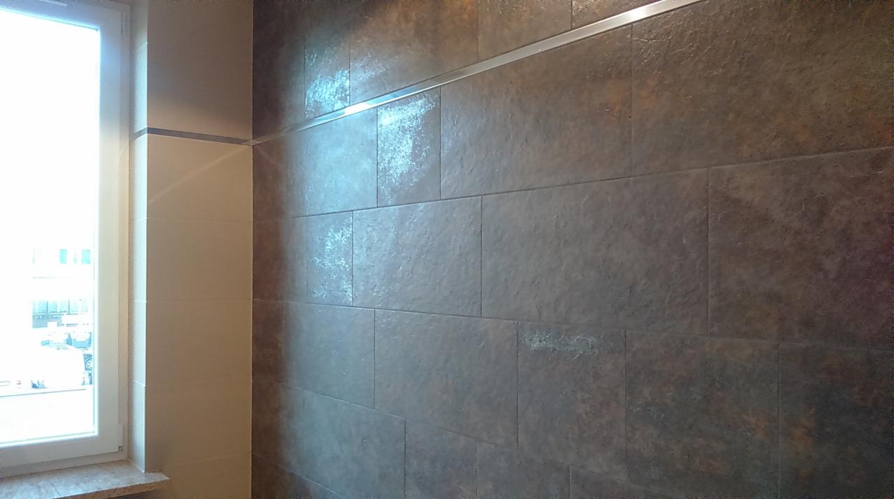 Wykończona ściana w łazience z brązowych płytek imitujących kamień, oddzielonych metalową listwą. Widoczne okno z białymi ramami i jasne płytki obok.