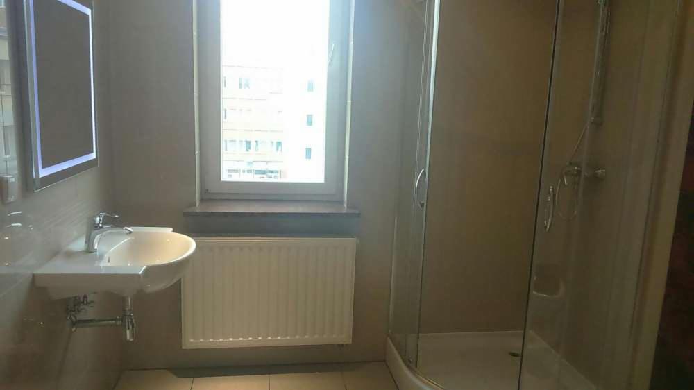 Jasna łazienka z kabiną prysznicową, umywalką i oknem z widokiem na budynek. Minimalistyczny design, neutralna kolorystyka ścian i podłogi, oświetlone lustro.