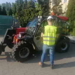 Operator w kamizelce odblaskowej i kasku stoi tyłem do kamery obok podnośnika teleskopowego Manitou z widłami na kostce brukowej, drzewa w tle.