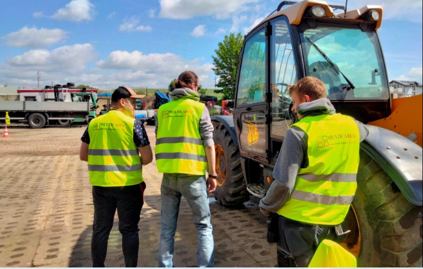 Trzech pracowników w żółtych kamizelkach odblaskowych z logo firmy stoi obok żółtej ładowarki teleskopowej JCB na placu budowy; w tle widoczne inne maszyny budowlane i błękitne niebo z chmurami.