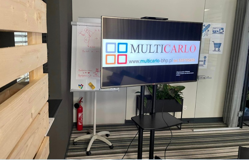 Sala szkoleniowa z ekranem prezentującym logo Multicarlo-bhp.pl, tablica suchościeralna z notatkami, gaśnica i rośliny doniczkowe w tle.