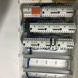 Zakład Mechaniczny Marek Kopala - Otwarta biała szafa rozdzielcza z zamontowanymi bezpiecznikami, przekaźnikami i listwami zaciskowymi, okablowanie w kolorze niebieskim i czarnym, widoczne szyny montażowe.