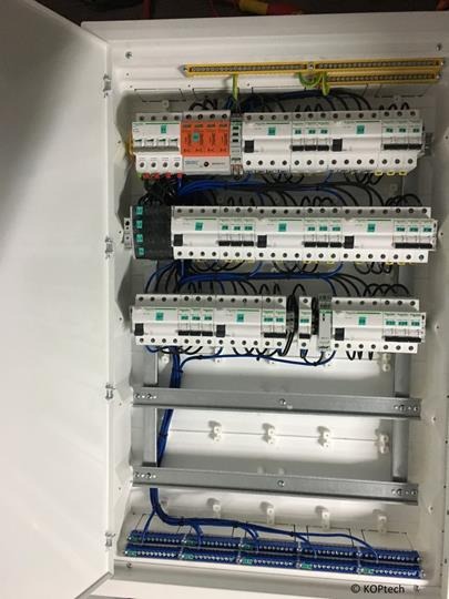 Otwarta biała szafa rozdzielcza z zamontowanymi bezpiecznikami, przekaźnikami i listwami zaciskowymi, okablowanie w kolorze niebieskim i czarnym, widoczne szyny montażowe.