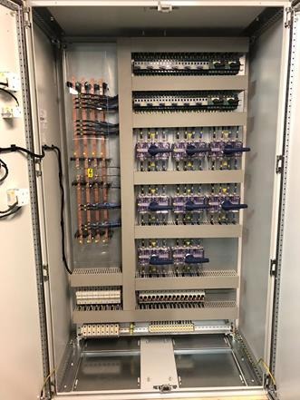 Otwarta szafa sterownicza z widocznymi przewodami, bezpiecznikami i innymi elementami instalacji elektrycznej, uporządkowane okablowanie.