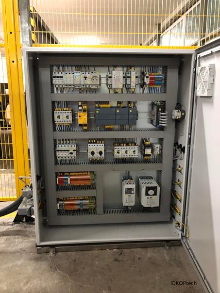 Otwarta szara szafa sterownicza z widocznymi licznymi elementami elektrycznymi, w tym przekaźnikami, bezpiecznikami i sterownikami PLC, z przewodami uporządkowanymi w listwach, na tle żółtej...