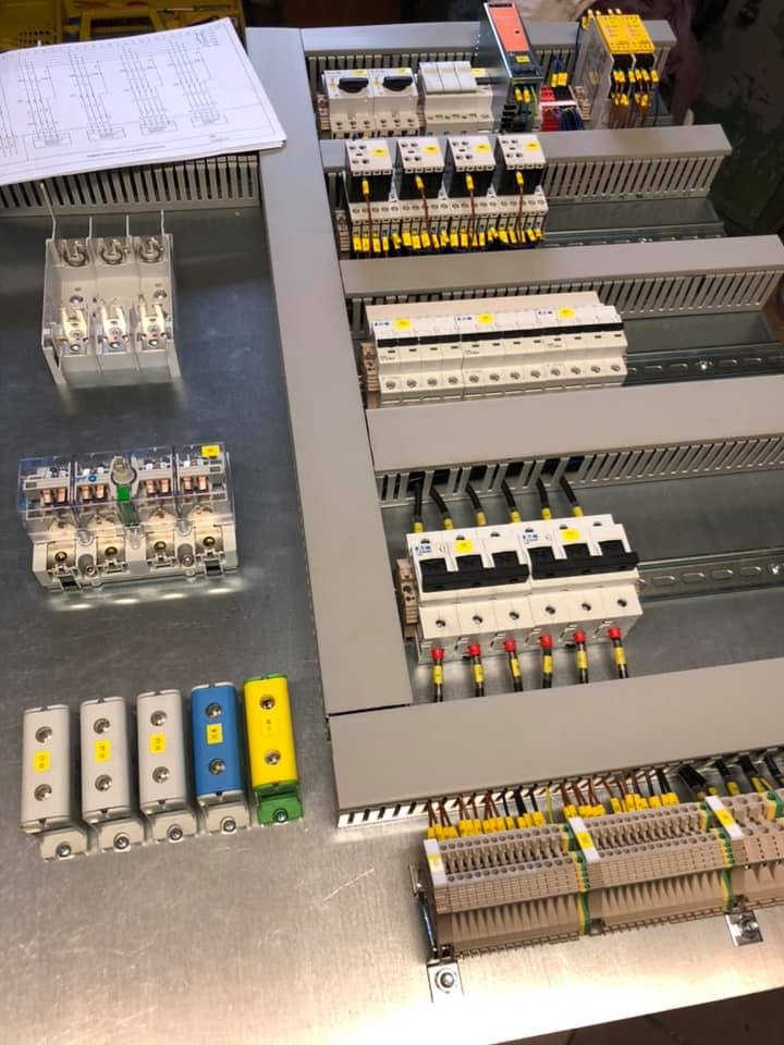 Rozdzielnica elektryczna w trakcie montażu, widoczne zabezpieczenia, styczniki, listwy zaciskowe, przewody z tulejkami oraz schemat ideowy na stole warsztatowym.