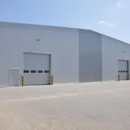 Nowoczesna hala przemysłowa z szarej blachy trapezowej, z dwoma bramami segmentowymi i drzwiami wejściowymi, zabezpieczona żółto-czarnymi słupkami odbojowymi, na tle kostki brukowej.