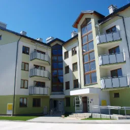 Nowoczesny budynek mieszkalny z balkonami i dużymi oknami, wykończony w kolorach bieli i pastelowej zieleni, widoczne wejście z podjazdem dla osób niepełnosprawnych.