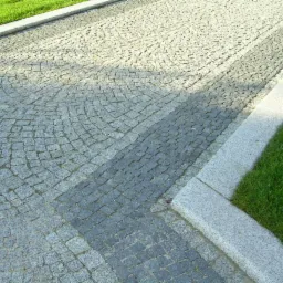 Ułożona z kostki granitowej ścieżka w ogrodzie, z widocznym wzorem łukowym i prostym, obrzeżem z kamienia oraz fragmentem trawnika. Różne odcienie szarości kostki.