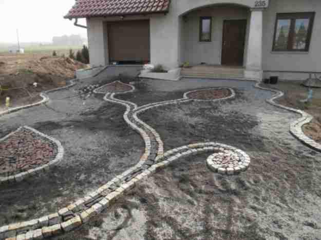 Ogród przed domem z dekoracyjną ścieżką z kostki, układającą się we wzory liści i spirali. Wypełnienia z drobnego żwiru w kształcie łez i koła. Dom z szarą elewacją w tle.