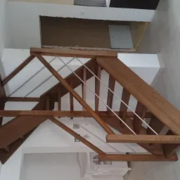 Konstrukcja schodów z jasnego drewna z metalowymi barierkami, widok z dołu, na tle białych ścian i jasnej podłogi wyłożonej płytkami.