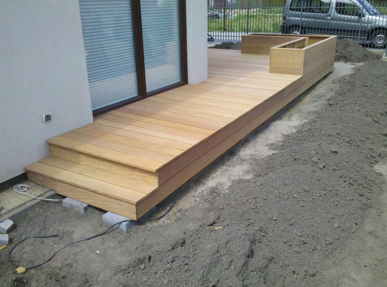 Nowo wybudowany taras z jasnego drewna z dwoma stopniami, prowadzący do drzwi balkonowych, z drewnianą donicą wzdłuż krawędzi, otoczony ziemią przed zagospodarowaniem.