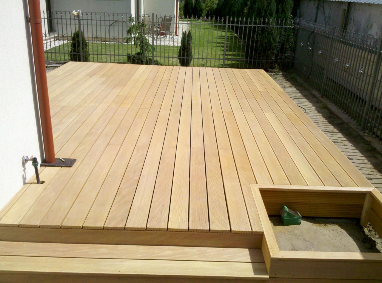 Jasne deski tworzą nowe podwyższenie tarasu z małym podestem i wbudowaną donicą z zieloną konewką w środku, na tle trawnika i metalowego ogrodzenia.