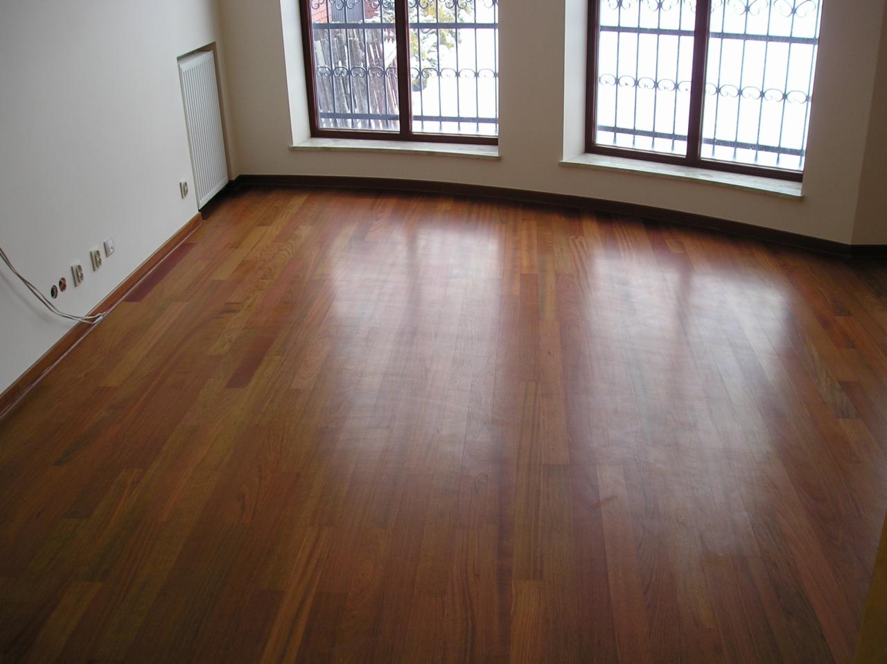 Wypolerowana, drewniana podłoga parkietowa w pustym pokoju z dwoma oknami zabezpieczonymi ozdobnymi kratami, widoczne gniazdka elektryczne przy listwie przypodłogowej.