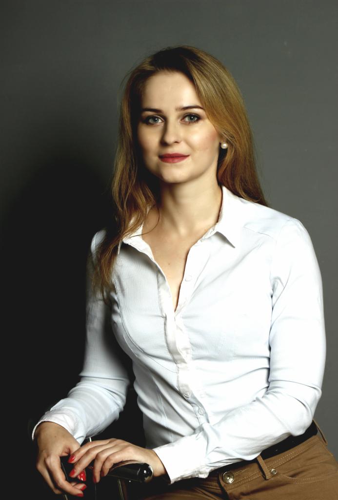 Portret kobiety z blond włosami w białej koszuli na szarym tle. Delikatny uśmiech, profesjonalny wygląd, subtelny makijaż. Częściowo widoczne ręce z czerwonym lakierem.