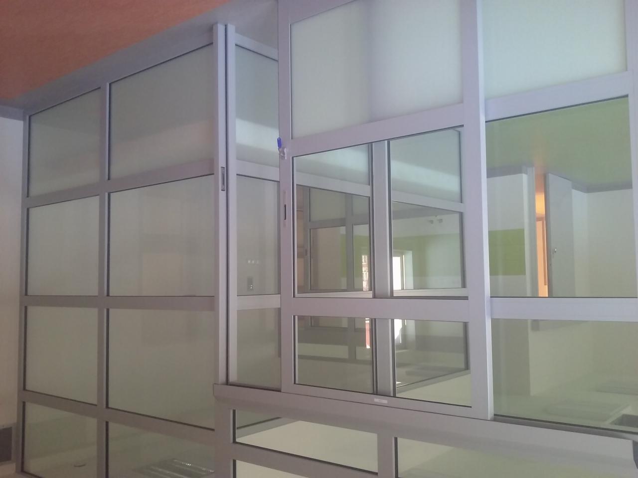 Szklana ścianka działowa z przesuwnymi oknami w aluminiowej ramie, widok z dołu, w tle widoczne wnętrze pomieszczenia.