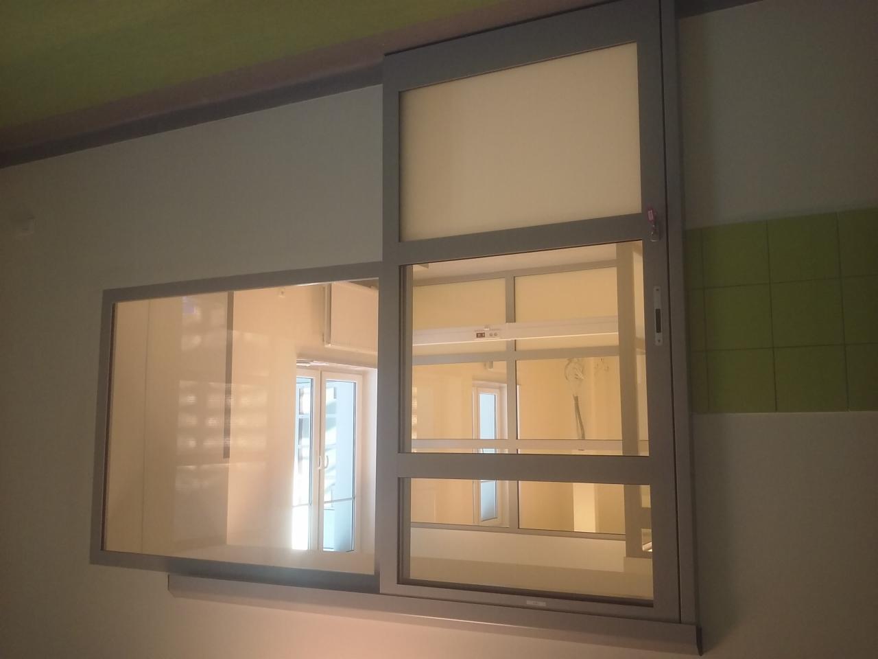 Przesuwne okno wewnętrzne z szarą ramą, widok na jasne pomieszczenie z oknem. Część okna zasłonięta mleczną szybą, ściana w tle z zielonymi płytkami.
