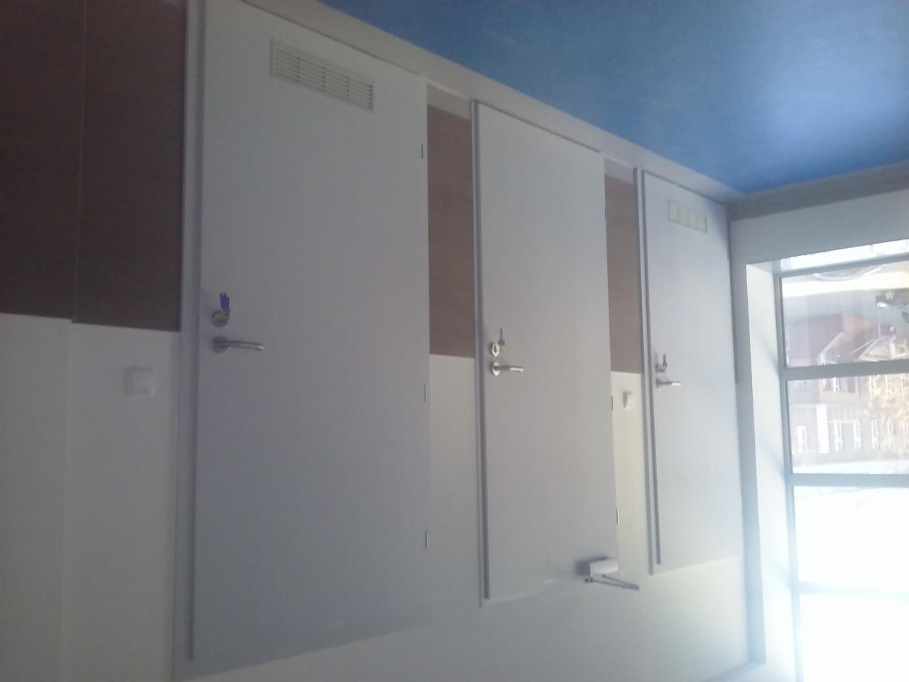 Trzy białe drzwi z metalowymi klamkami w publicznej toalecie, widoczne fragmenty ścian i okno z widokiem na zewnątrz, sufit pomalowany na niebiesko.