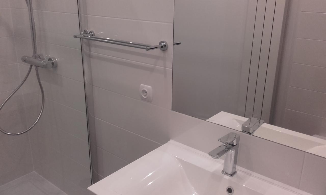 Nowoczesna, minimalistyczna łazienka z białymi płytkami, chromowaną armaturą, szklanym panelem prysznicowym i grzejnikiem drabinkowym.