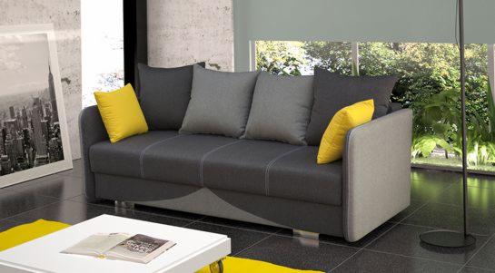 Rozkładana sofa w odcieniach szarości z żółtymi poduszkami, ustawiona w nowoczesnym wnętrzu z widokiem na zieleń za oknem.