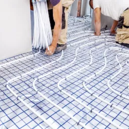 Białe rurki ogrzewania podłogowego układane na folii z nadrukowaną siatką w niebieskie kwadraty przez dwóch instalatorów w beżowych spodniach roboczych.