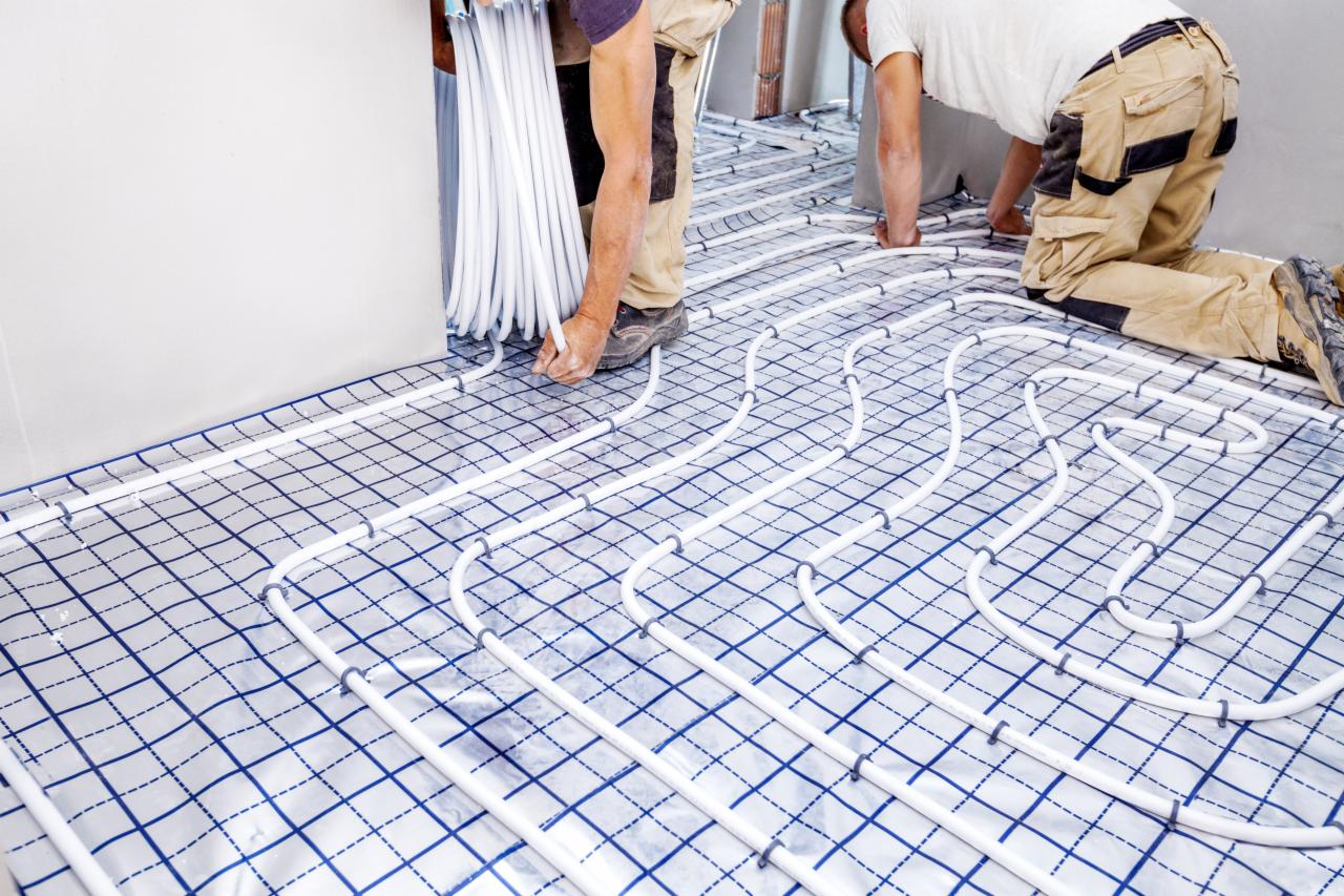 Białe rurki ogrzewania podłogowego układane na folii z nadrukowaną siatką w niebieskie kwadraty przez dwóch instalatorów w beżowych spodniach roboczych.