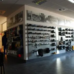 Jasna sala sprzedaży z ekspozycją obuwia roboczego, kasków ochronnych i akcesoriów na perforowanych ścianach, uzupełniona czarno-białymi fotografiami i dwoma krzesłami.