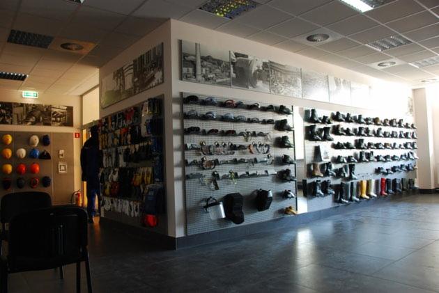 Jasna sala sprzedaży z ekspozycją obuwia roboczego, kasków ochronnych i akcesoriów na perforowanych ścianach, uzupełniona czarno-białymi fotografiami i dwoma krzesłami.