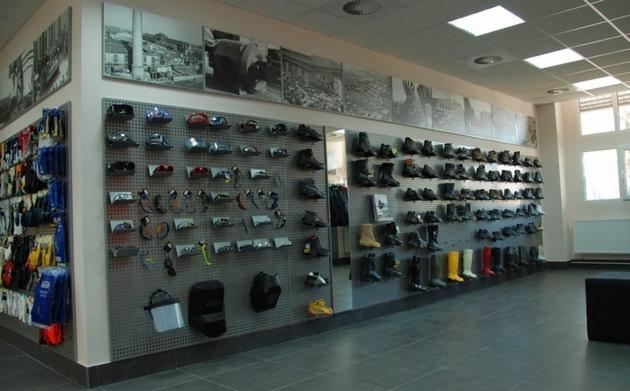 Wnętrze sklepu z artykułami BHP: ściana z ekspozycją obuwia roboczego, kasków, okularów ochronnych i akcesoriów, nad nią czarno-białe zdjęcia przemysłowe.