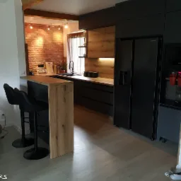 Nowoczesna kuchnia z czarnymi frontami szafek, drewnianymi akcentami i ceglaną ścianą w tle, widok na wyspę kuchenną z dwoma czarnymi hokerami.