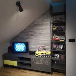 Szara szafka RTV z żółtym akcentem i drewnopodobnym frontem, ustawiona pod skosem dachu, obok metalowy regał z półkami, dekoracje i zabawki Lego.