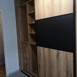 Wnętrze szafy przesuwnej z drewnopodobnymi frontami w odcieniach beżu i czerni, widoczne półki, drążek i szuflady, nowoczesny design.