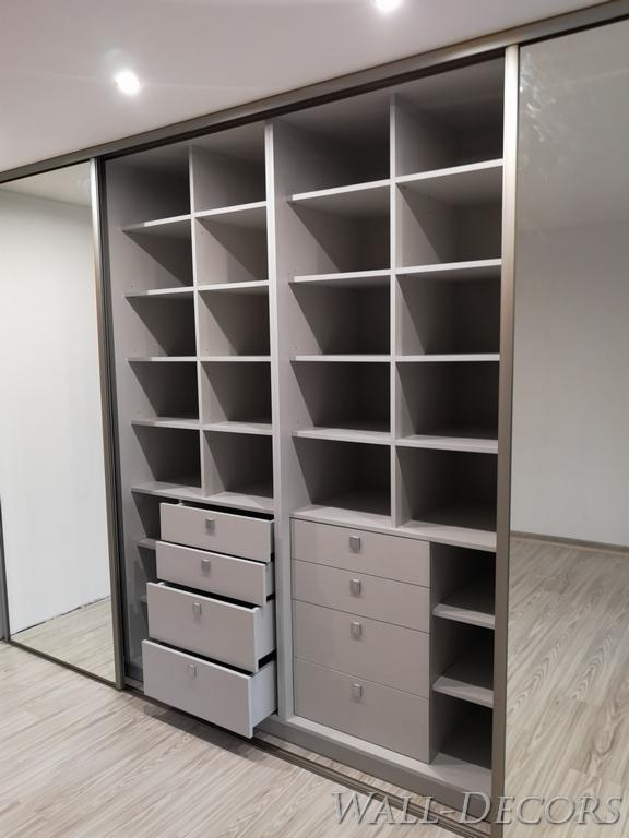 Jasnoszara szafa wnękowa z przesuwnymi drzwiami lustrzanymi, z otwartymi półkami i szufladami, z których jedna jest wysunięta.