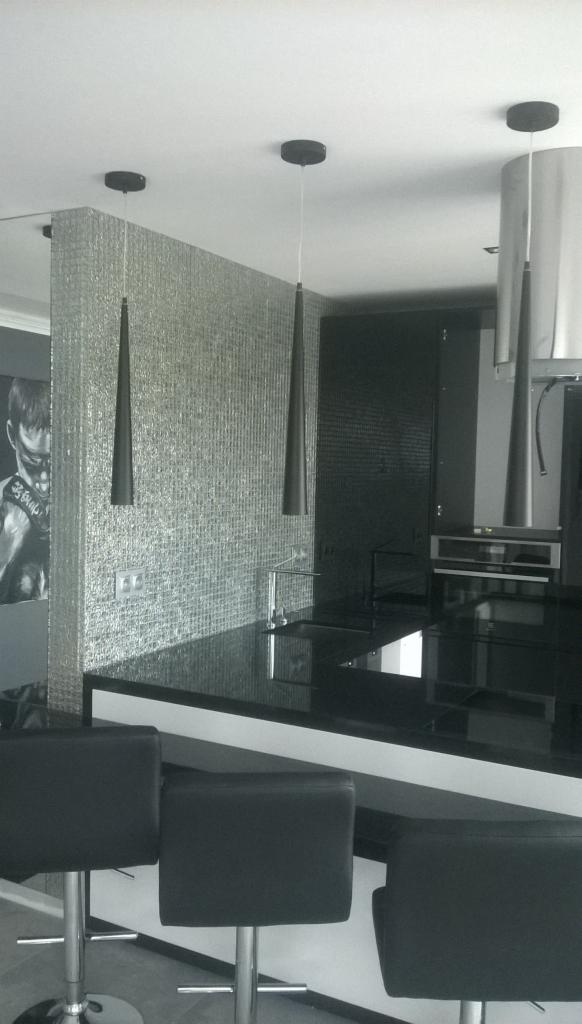 Nowoczesna kuchnia w odcieniach szarości z błyszczącą mozaiką na ścianie, czarnym blatem i trzema minimalistycznymi lampami wiszącymi, oraz czarnymi krzesłami barowymi. Na ścianie widoczna grafika...