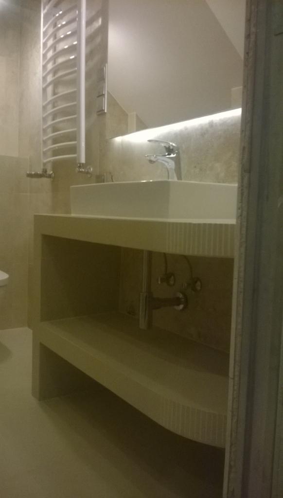 Nowoczesna łazienka z prostokątną umywalką nablatową, podświetlanym lustrem i grzejnikiem drabinkowym, wykończona w jasnych, beżowych tonacjach z widoczną instalacją hydrauliczną pod umywalką.