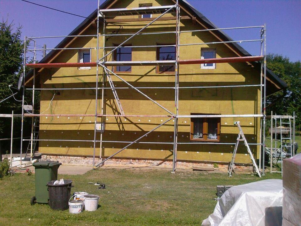 Dom w trakcie ocieplania, widoczne rusztowania i warstwa izolacyjna na elewacji. Detal: kwiaty w oknie na parterze.