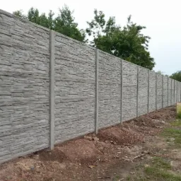 Długi, szary ogrodzenie betonowe o fakturze kamienia, z widocznymi słupkami, otoczone ziemią i zielenią. Tło stanowią drzewa i pochmurne niebo.