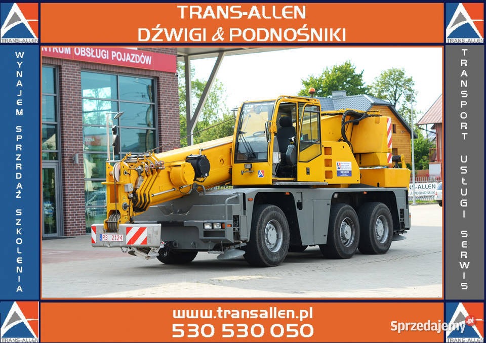 Żółty, samojezdny dźwig teleskopowy z wysięgnikiem, zaparkowany przed budynkiem serwisu pojazdów, widok z boku.