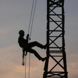 Sylwetka pracownika w kasku, opuszczającego się na linie z metalowej konstrukcji wieży na tle zachodzącego słońca.
