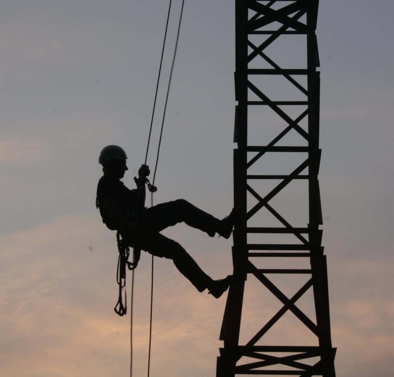 Sylwetka pracownika w kasku, opuszczającego się na linie z metalowej konstrukcji wieży na tle zachodzącego słońca.