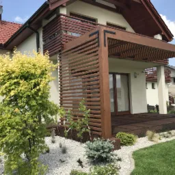 Nowoczesna drewniana pergola z tarasem, zintegrowana z budynkiem, otoczona białymi kamieniami i zielenią.