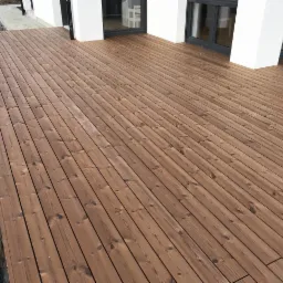 Świeżo położony taras drewniany z widocznymi słojami drewna, przylegający do białej elewacji budynku z ciemnymi oknami, ziemia wokół jeszcze niezagospodarowana.