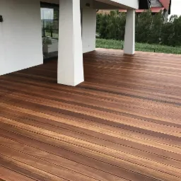 Nowoczesny taras z desek w odcieniach brązu, z widocznymi białymi filarami domu i zielenią w tle.