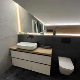 Nowoczesna łazienka z szarą płytką imitującą kamień, białą umywalką nablatową na szafce z drewnianym blatem, podświetlanym lustrem i toaletą w zabudowie.