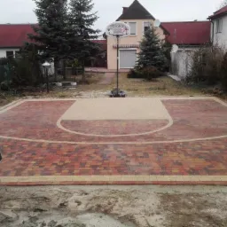 Boisko do koszykówki z kostki brukowej w Krosno Odrzańskie. Czerwono-żółta nawierzchnia, w tle domy i drzewa, betoniarka z lewej strony. Nietypowe zastosowanie.
