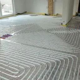 Białe rury grzewcze ułożone spiralnie na siatce podłogowej w jasnym pomieszczeniu w trakcie instalacji ogrzewania podłogowego, widoczne połączenia rur w kolorze niebieskim i czerwonym.